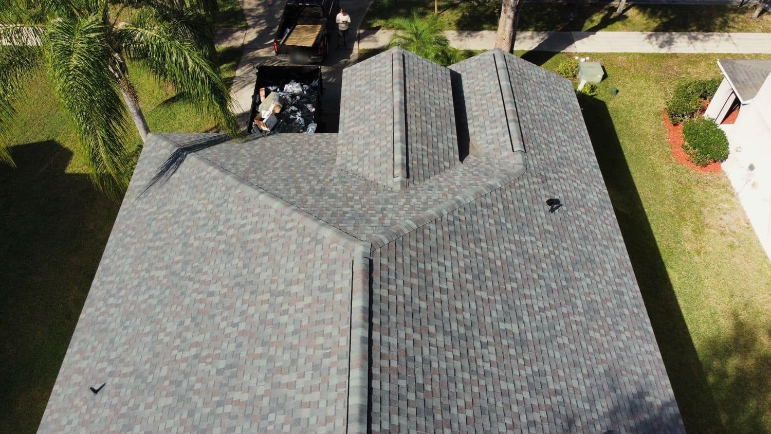 Drone view looking down on a gray-blend shingle roof with two front gables, a debris container and a pickup in the driveway beyond.