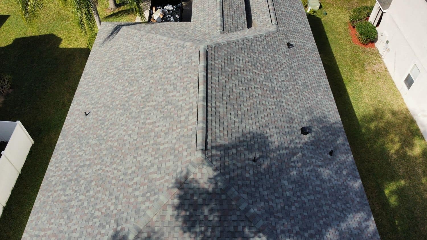 Top-down drone view of a gray-blend shingle roof with ridges, valleys and roof vents, lawn on both sides and a white fence at the left edge.