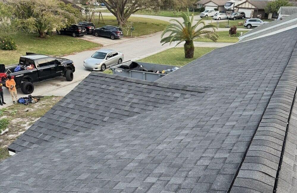 View from a roof ridge over dark gray architectural shingles toward the street, with two workers beside a black pickup and a gray roll-off container at the curb.