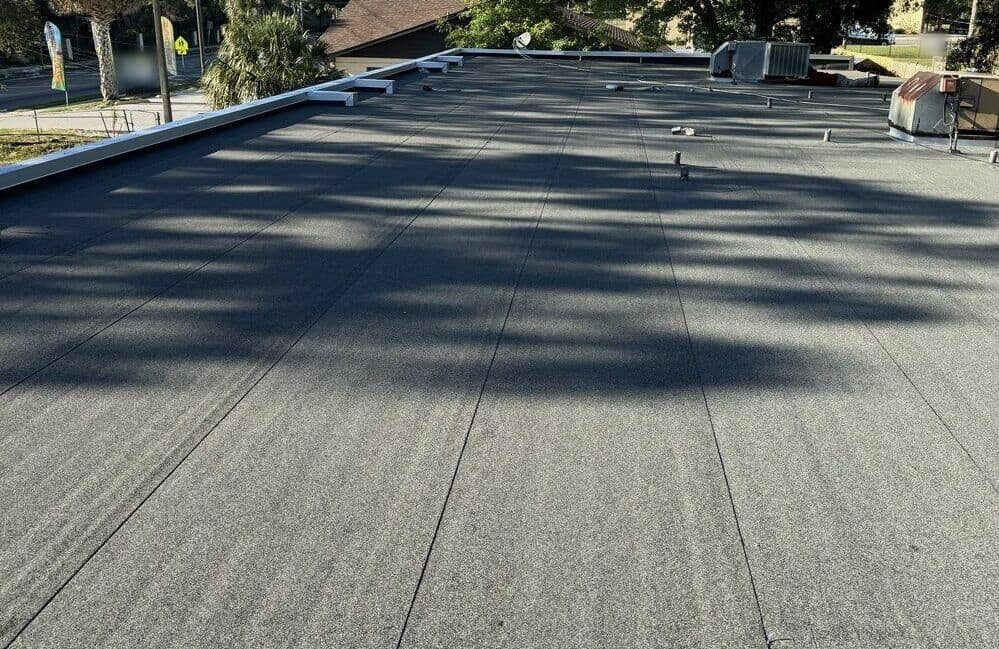 A flat roof covered in a gray granulated cap sheet, with rooftop units and vents in the distance and tree shadows across the surface.