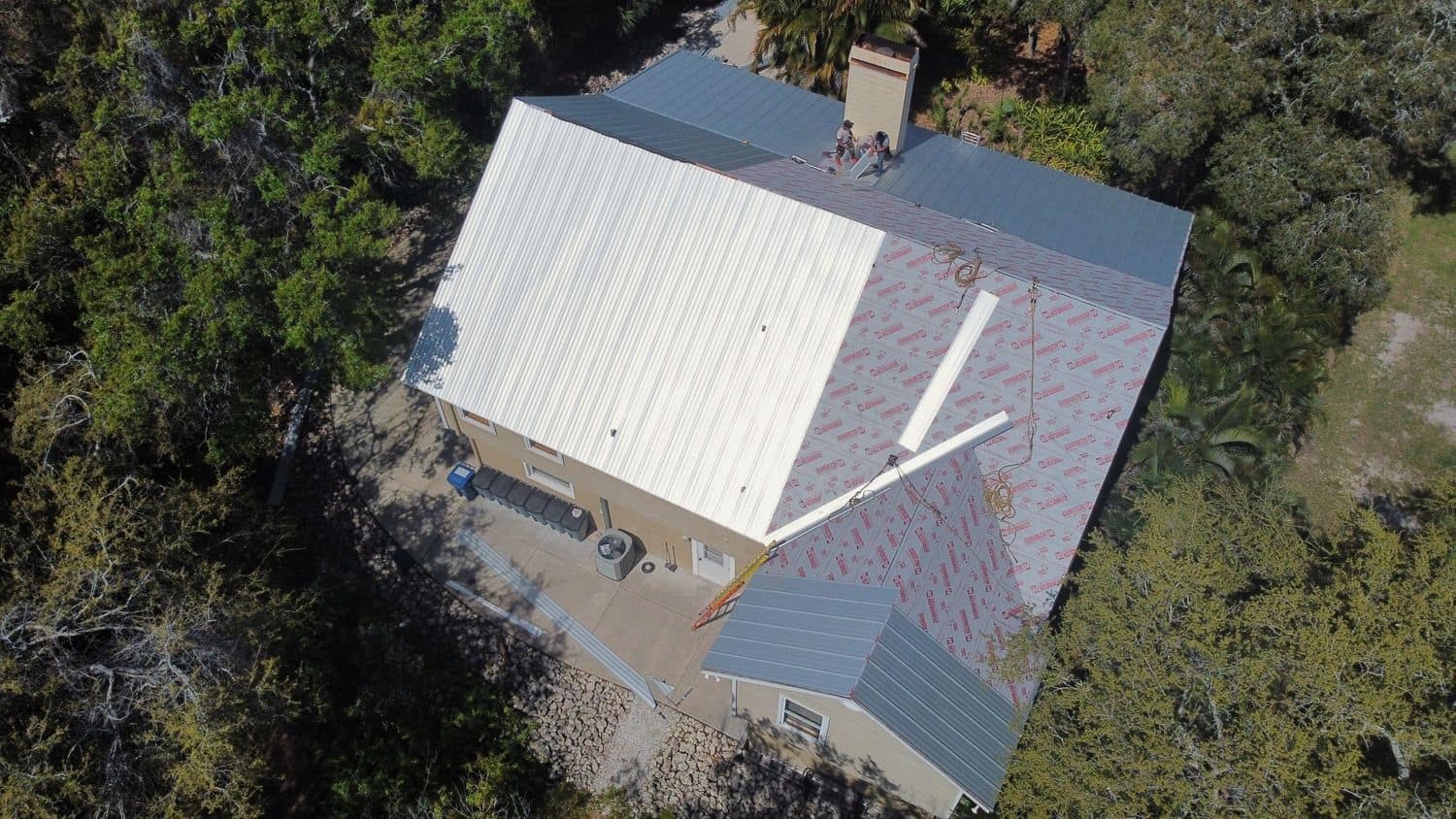 Top-down drone view of a home with silver metal roof panels partly installed over red-printed synthetic underlayment, two workers near the chimney and woods all around.