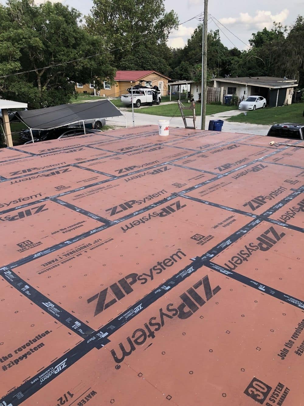 A roof deck sheathed in red-brown ZIP System panels with black seam tape, a bucket and a ladder on the deck, homes and a street behind.
