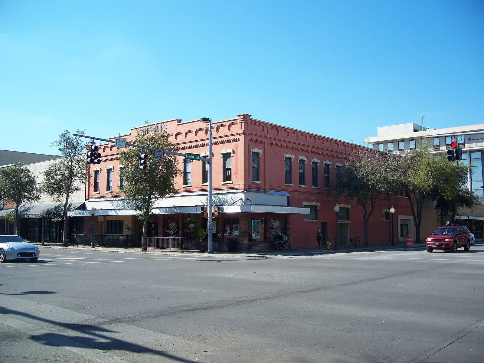 A red-brick historic corner building with awnings stands at a sunny intersection with traffic signals and parked cars.