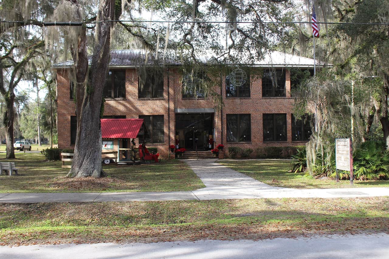Two-story brick town hall with a metal roof, draped in Spanish moss, with a walkway and landscaped lawn.