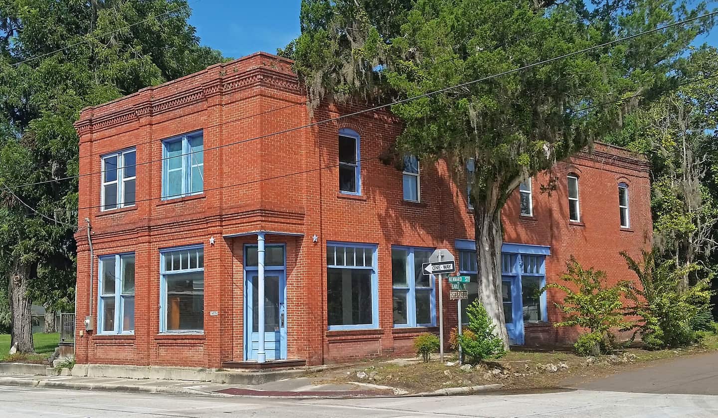 Two-story red brick corner building with blue-trimmed windows and doors, shaded by a large oak tree.