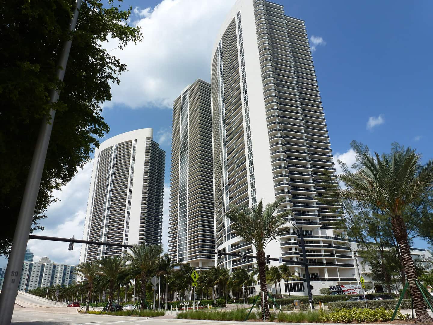 Three tall white high-rise residential towers under blue sky with palm trees along a landscaped street.