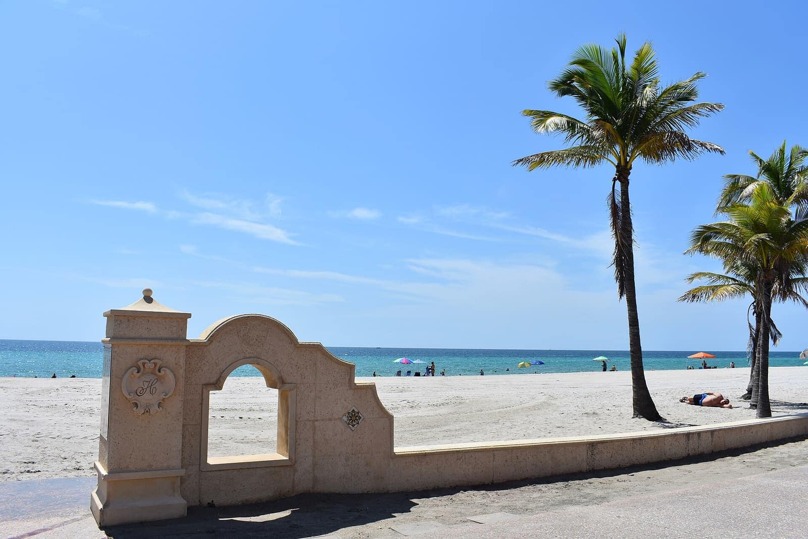 Ornamental beige beach wall with an arched opening, palm trees, and blue ocean under bright sky.