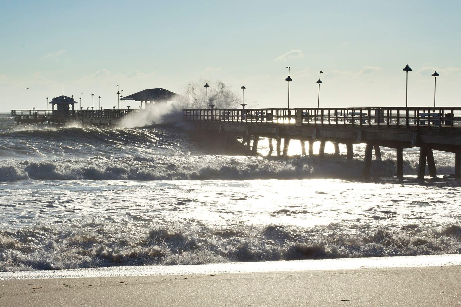 Waves crash beside a long fishing pier under bright sunlight with a pavilion at the far end.