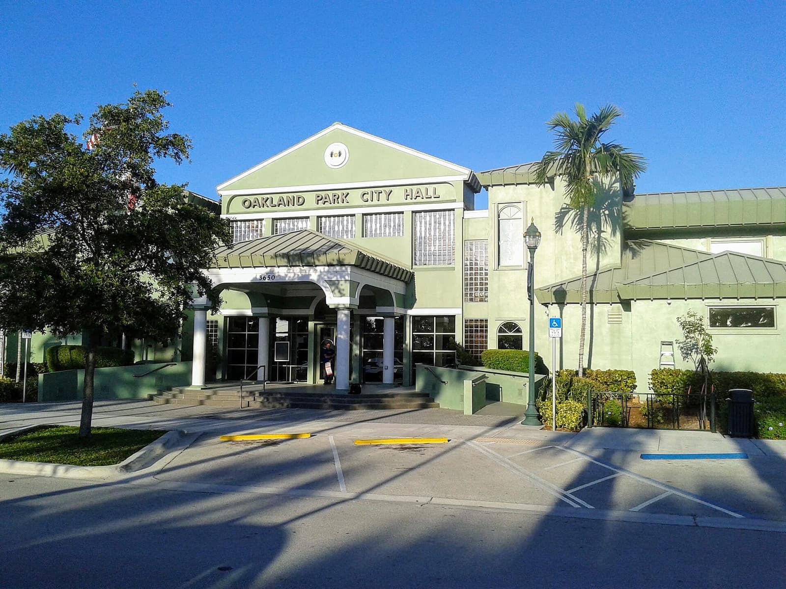 Green two-story city hall building with columned entrance and palm trees under clear blue sky.