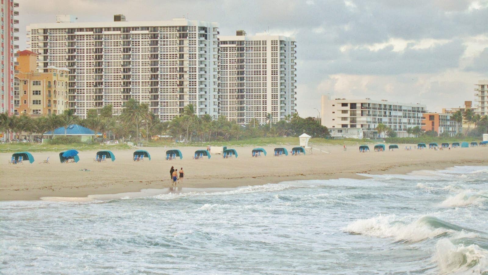 Sandy beach with blue cabanas, tall white condo towers, and two distant people walking at the shoreline.