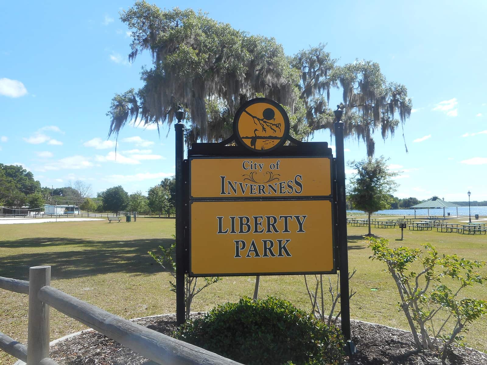 City of Inverness Liberty Park sign with moss-draped trees, a grassy lawn, and a lake in the distance.