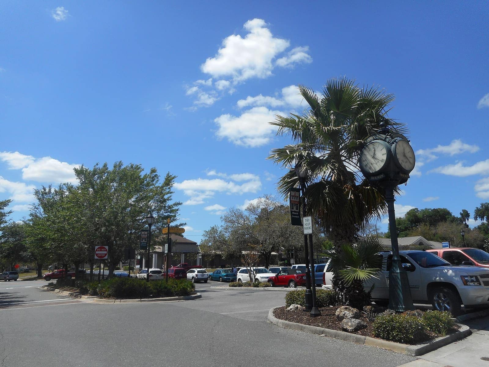 Ornate street clock on a post beside a palm tree, with parked cars and a sunny blue sky.