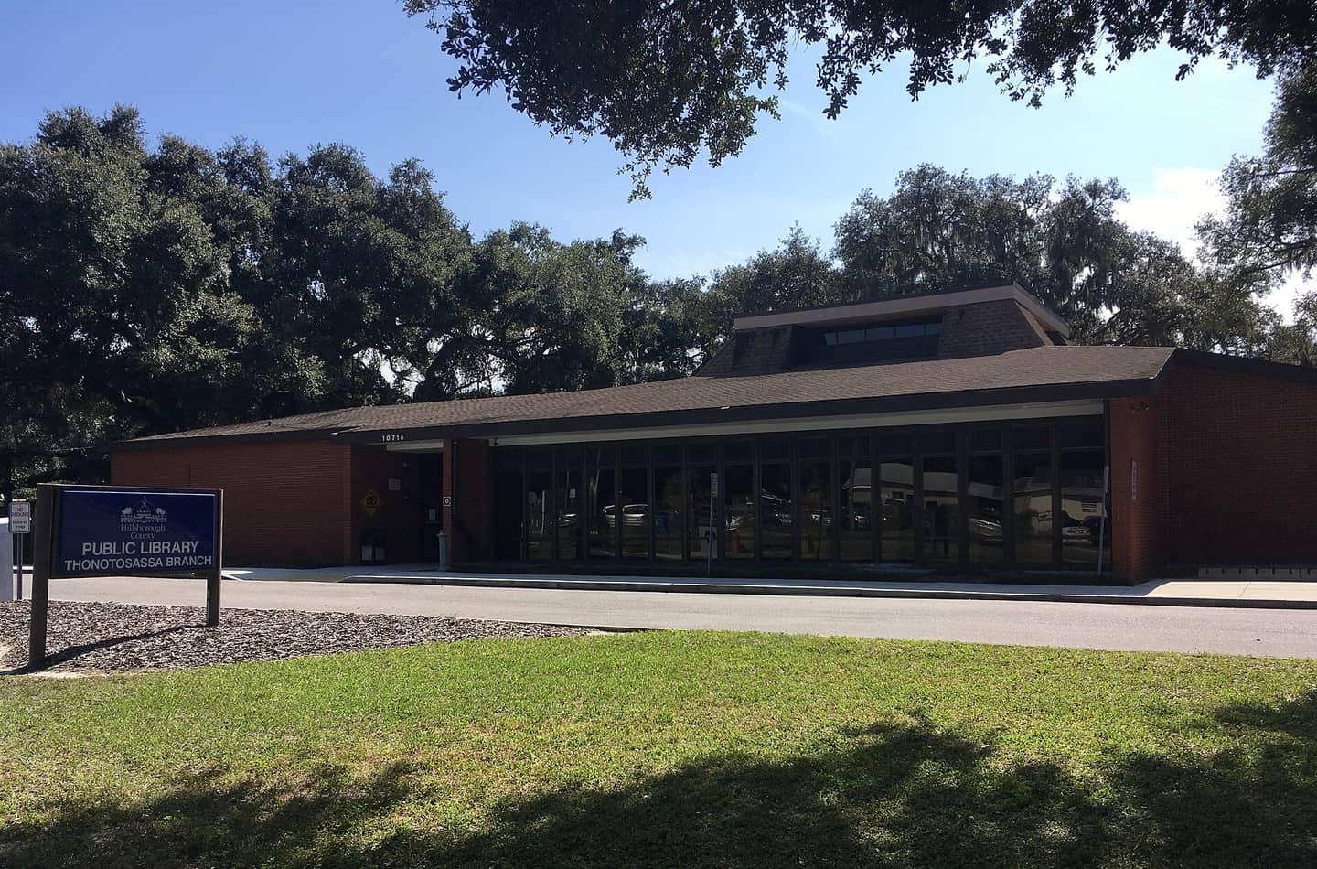 Single-storey brick library building with a dark roof behind a blue entrance sign and oak trees.