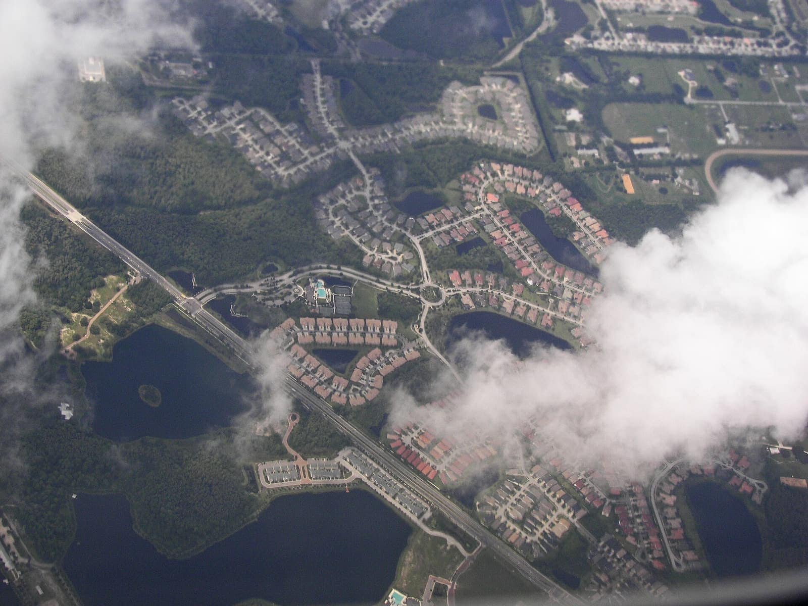 Aerial view of a suburban neighborhood with curving streets, a traffic circle, lakes and a highway through wooded land.