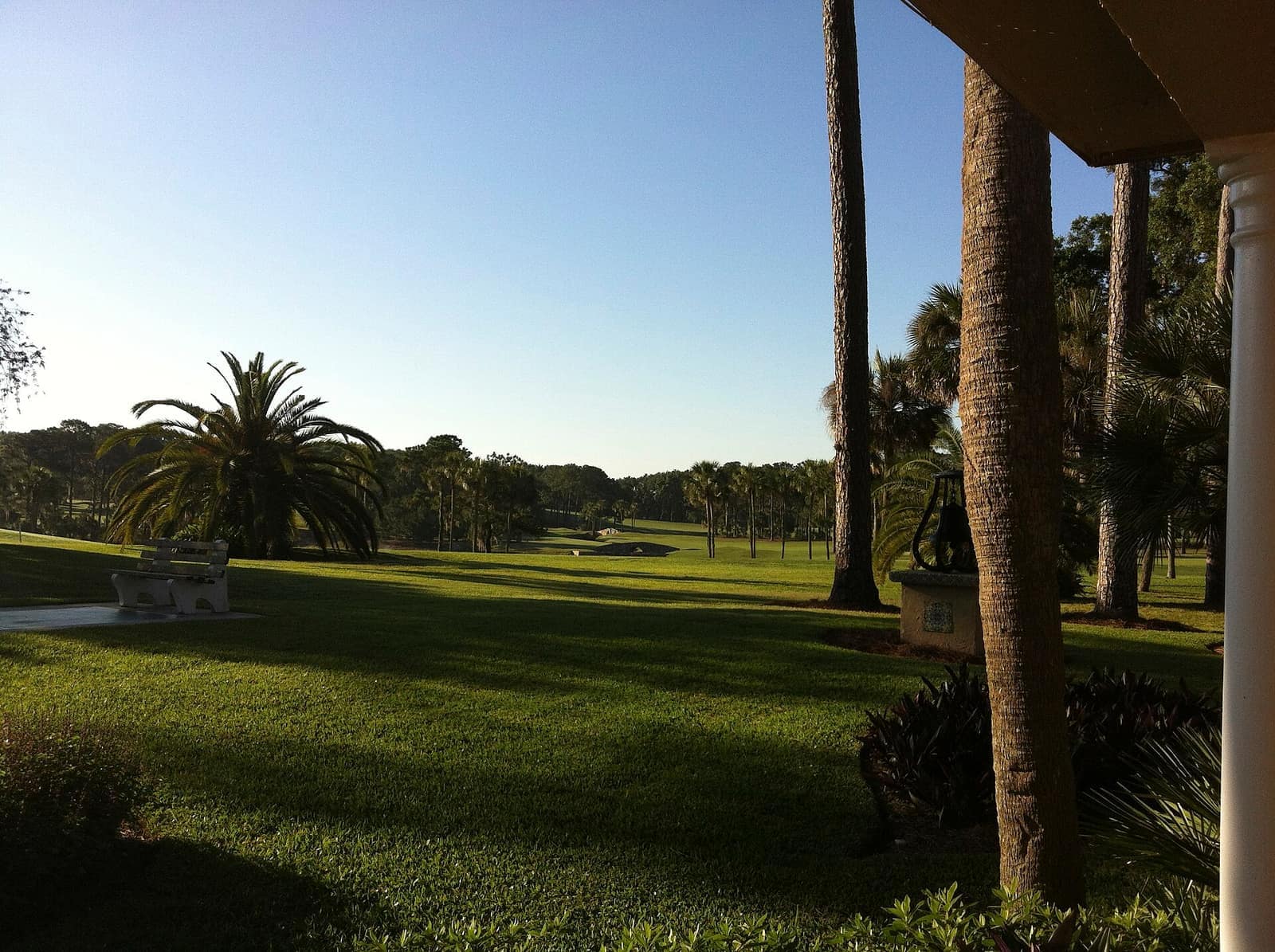 Green golf fairway lined with palm trees, a stone bench and a columned porch under a clear sky.