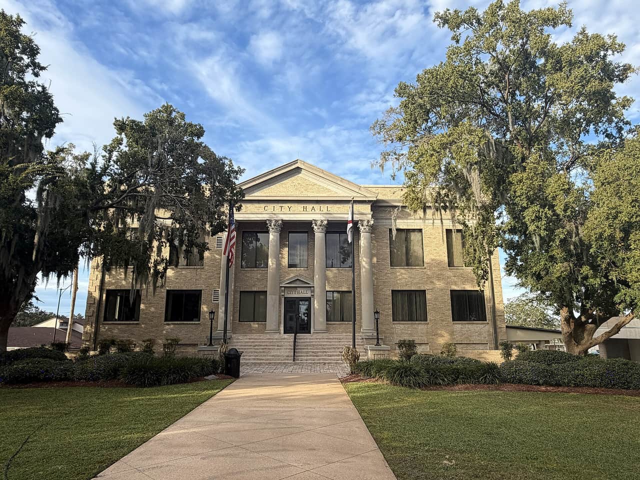 Two-story tan brick city hall with columns, framed by live oaks and a blue sky.