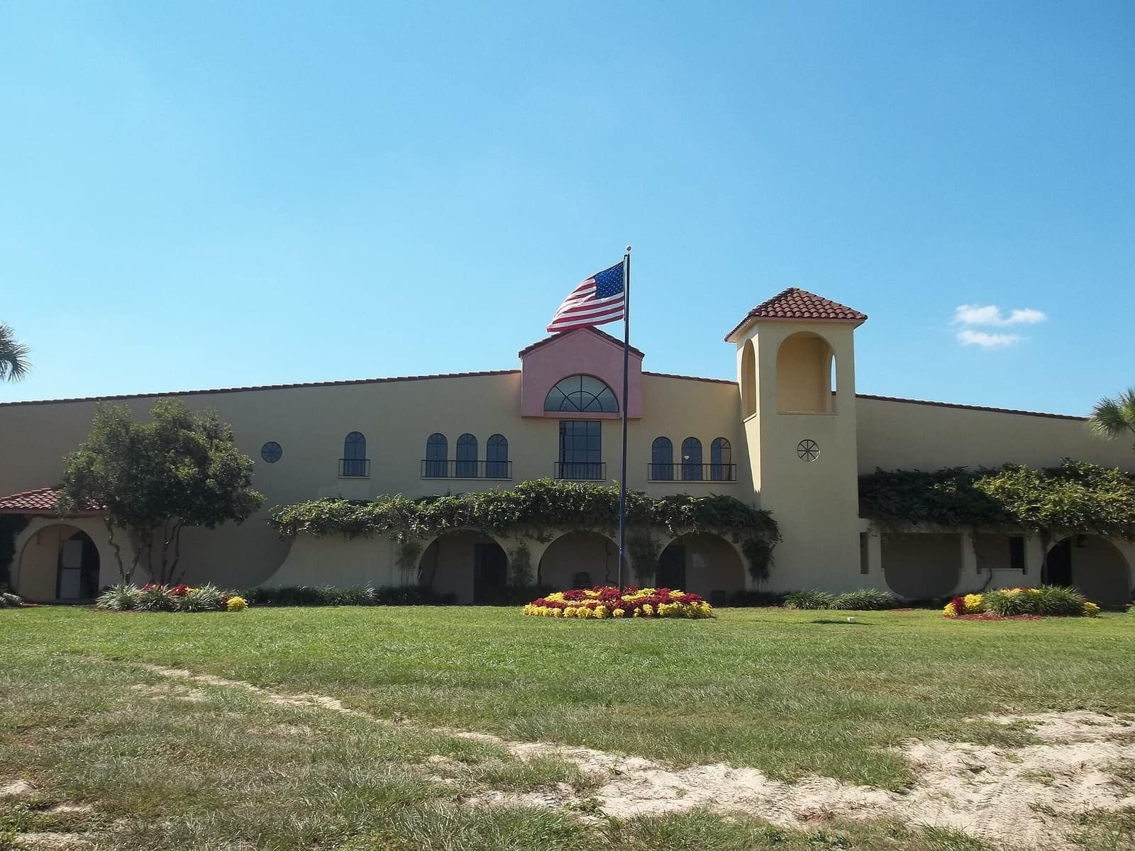 Cream stucco winery building with red tile roof, American flag, arched windows, lawn and flower beds in daylight.