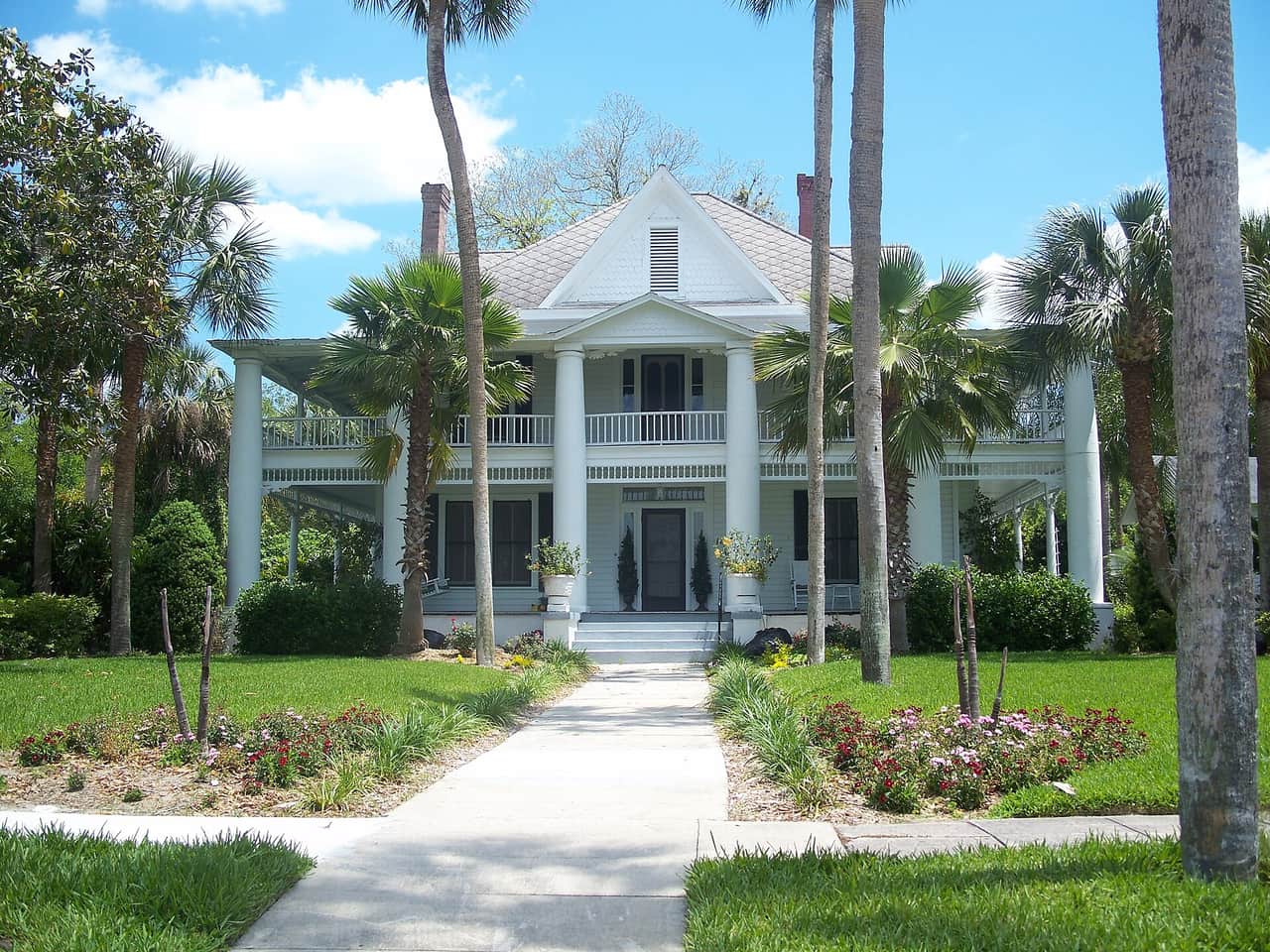 White two-story colonial-style house with columned porches, palm trees and a walkway through the lawn.