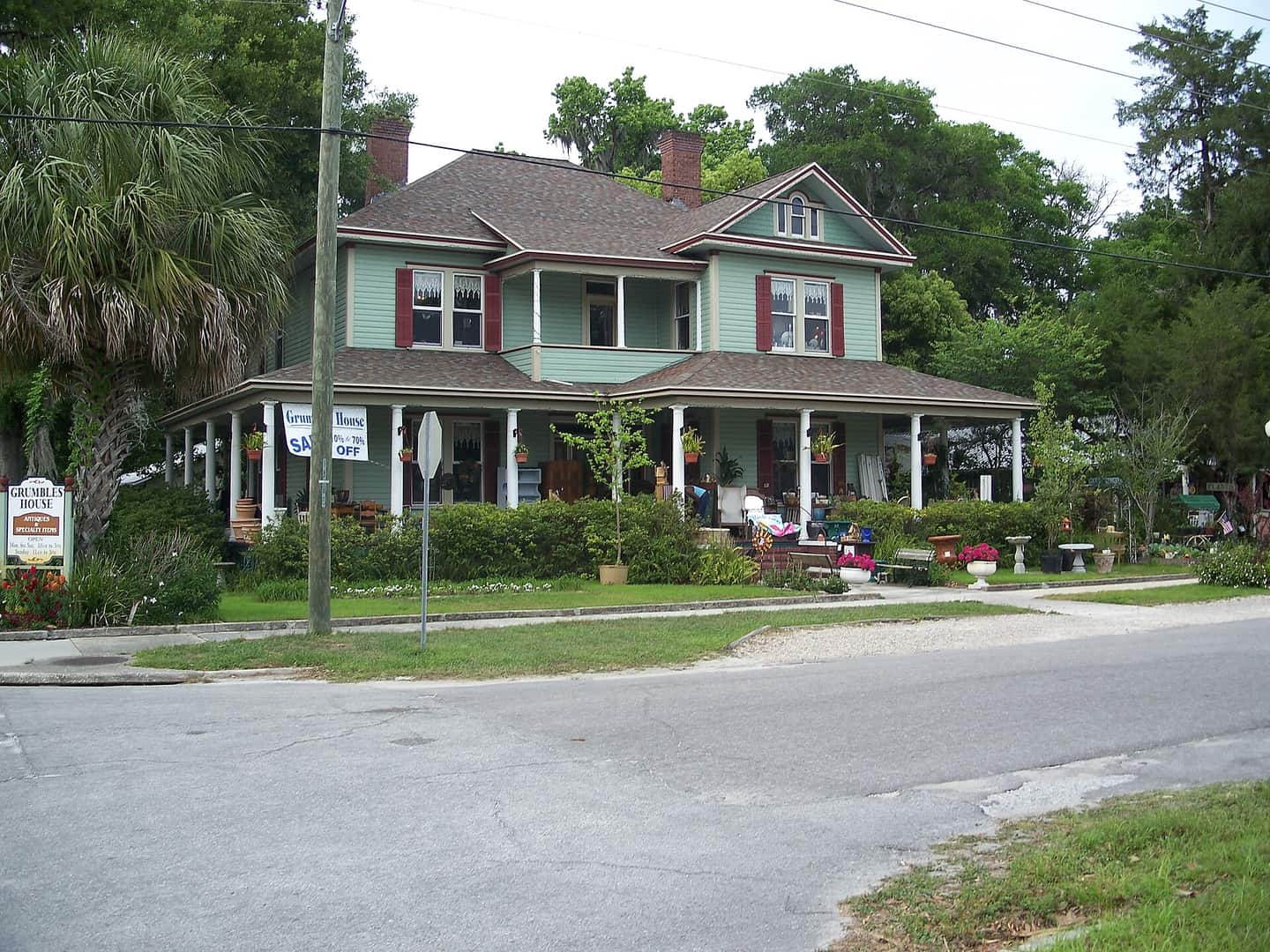 Historic two-storey house with a long front porch, white columns and shrubs beside a tree-lined street.