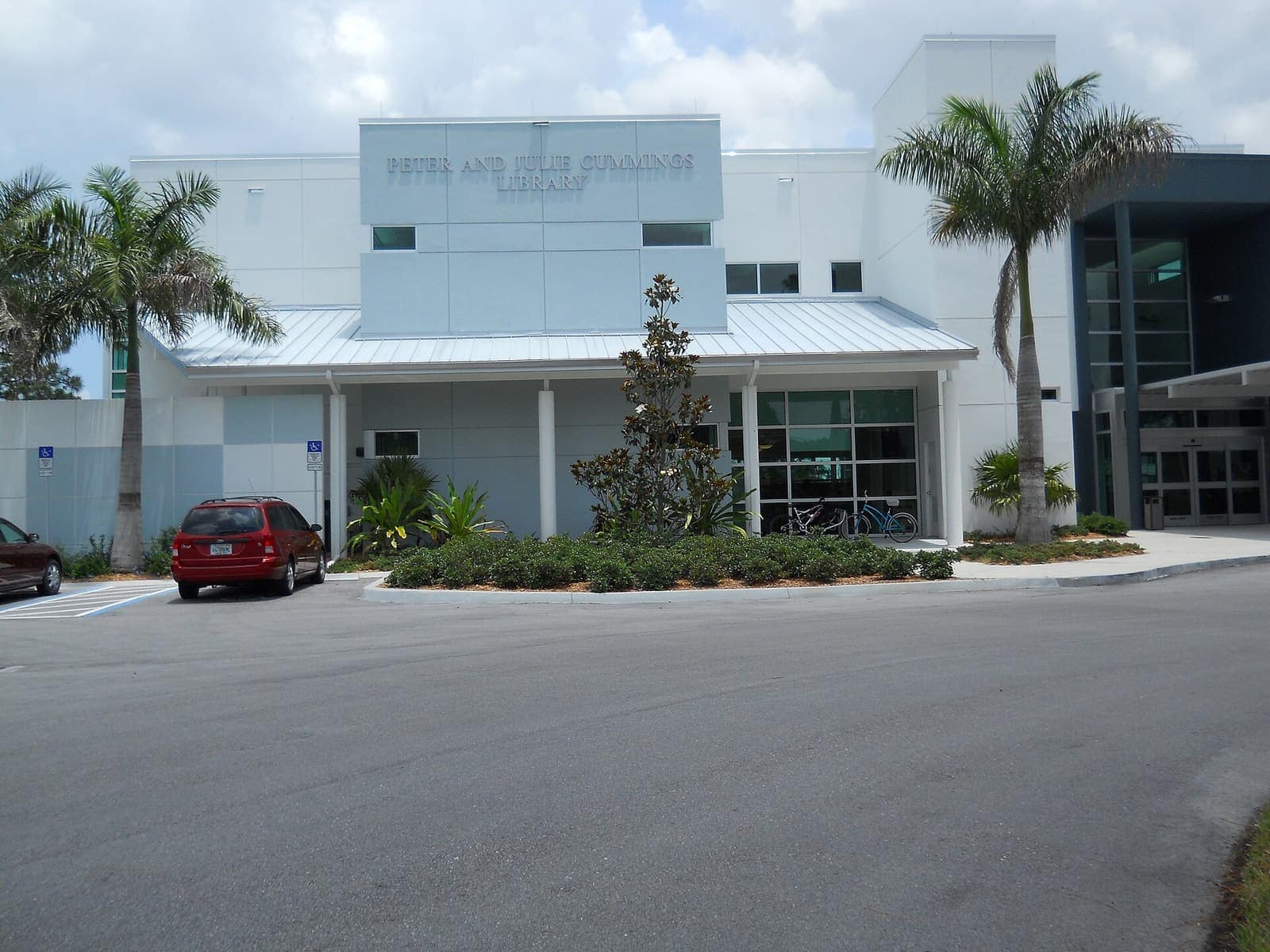White modern library building with a metal-roofed entrance, palm trees, and a parked red minivan.