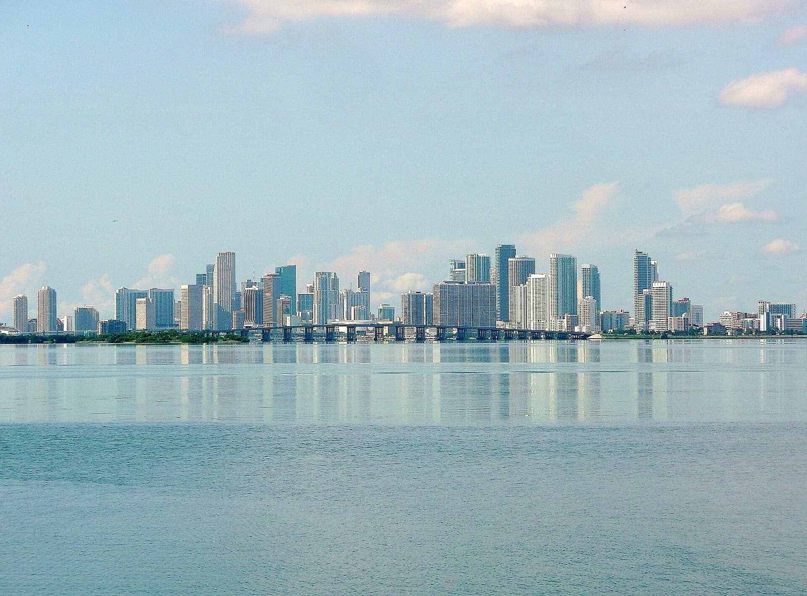 Miami city skyline of high-rise towers and a causeway bridge across calm bay water under pale sky.