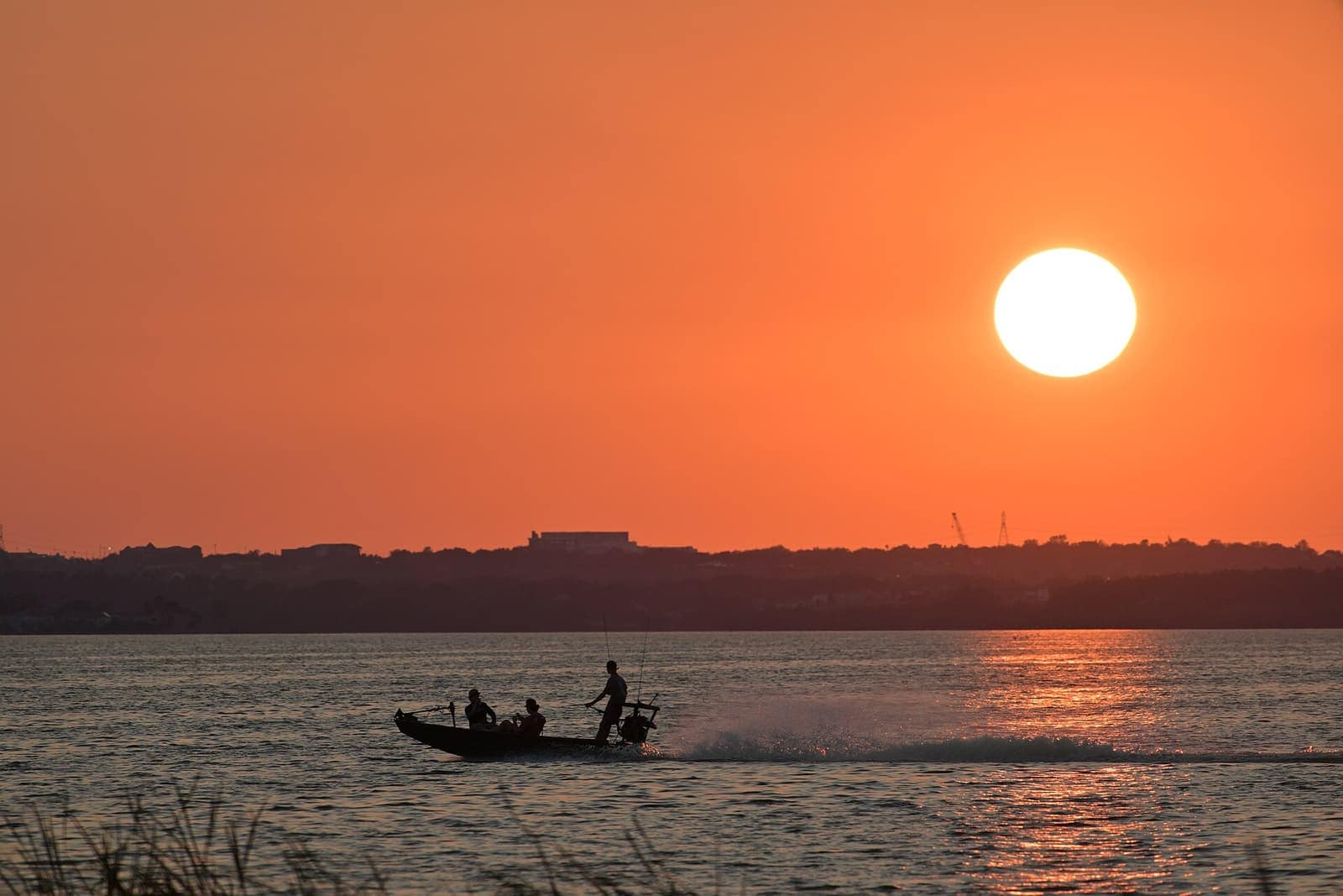 Orange sunset over a lake with a small boat and silhouetted people, distant shoreline beyond.