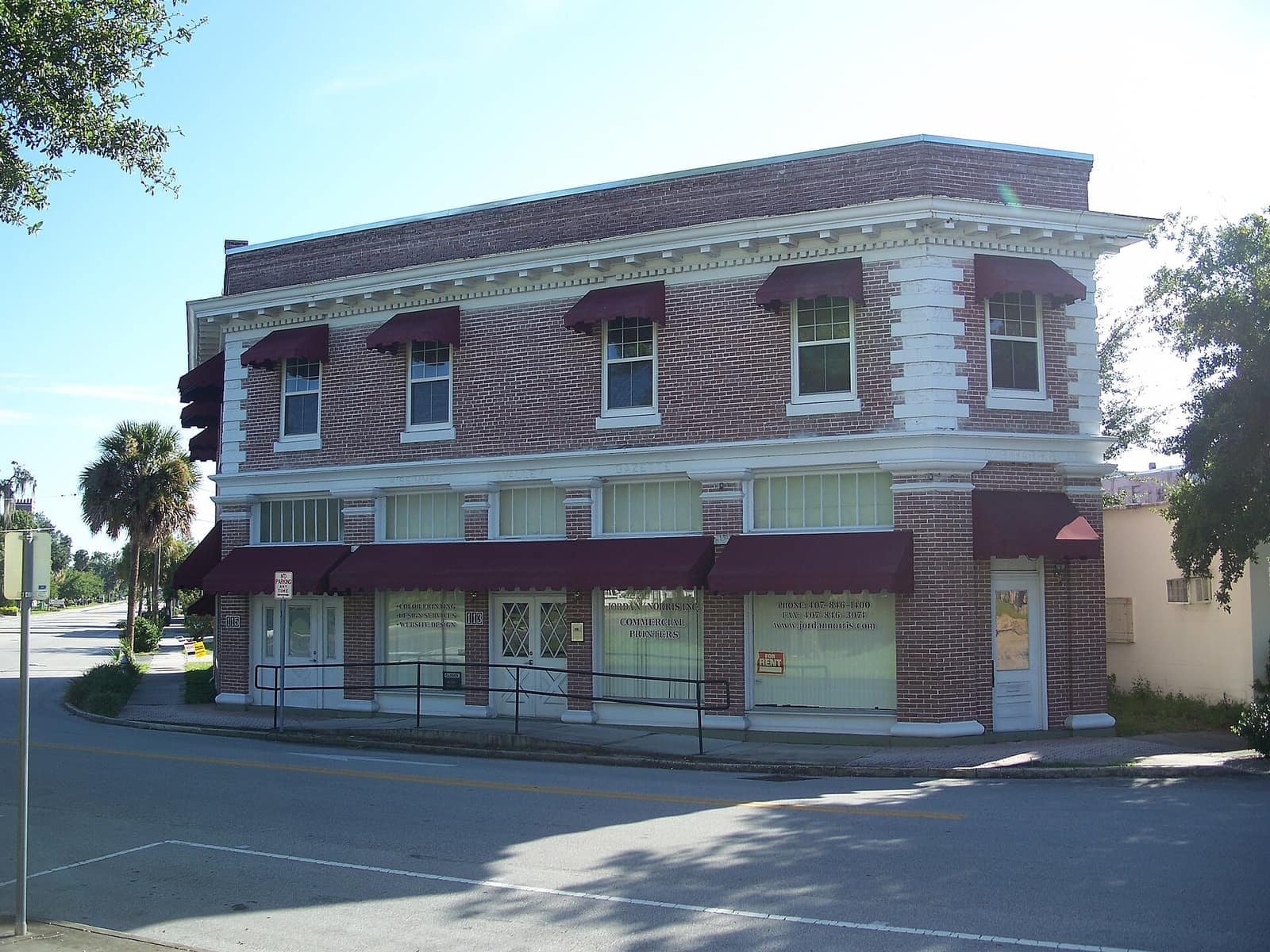 Two-story brick historic commercial building on a street corner with red awnings under blue sky.