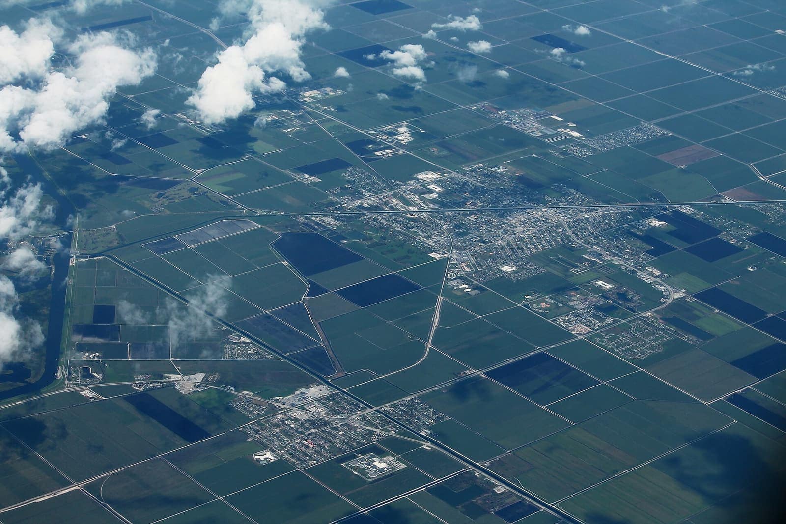 Aerial view of a town surrounded by flat green farm fields, with scattered clouds and grid-like roads.
