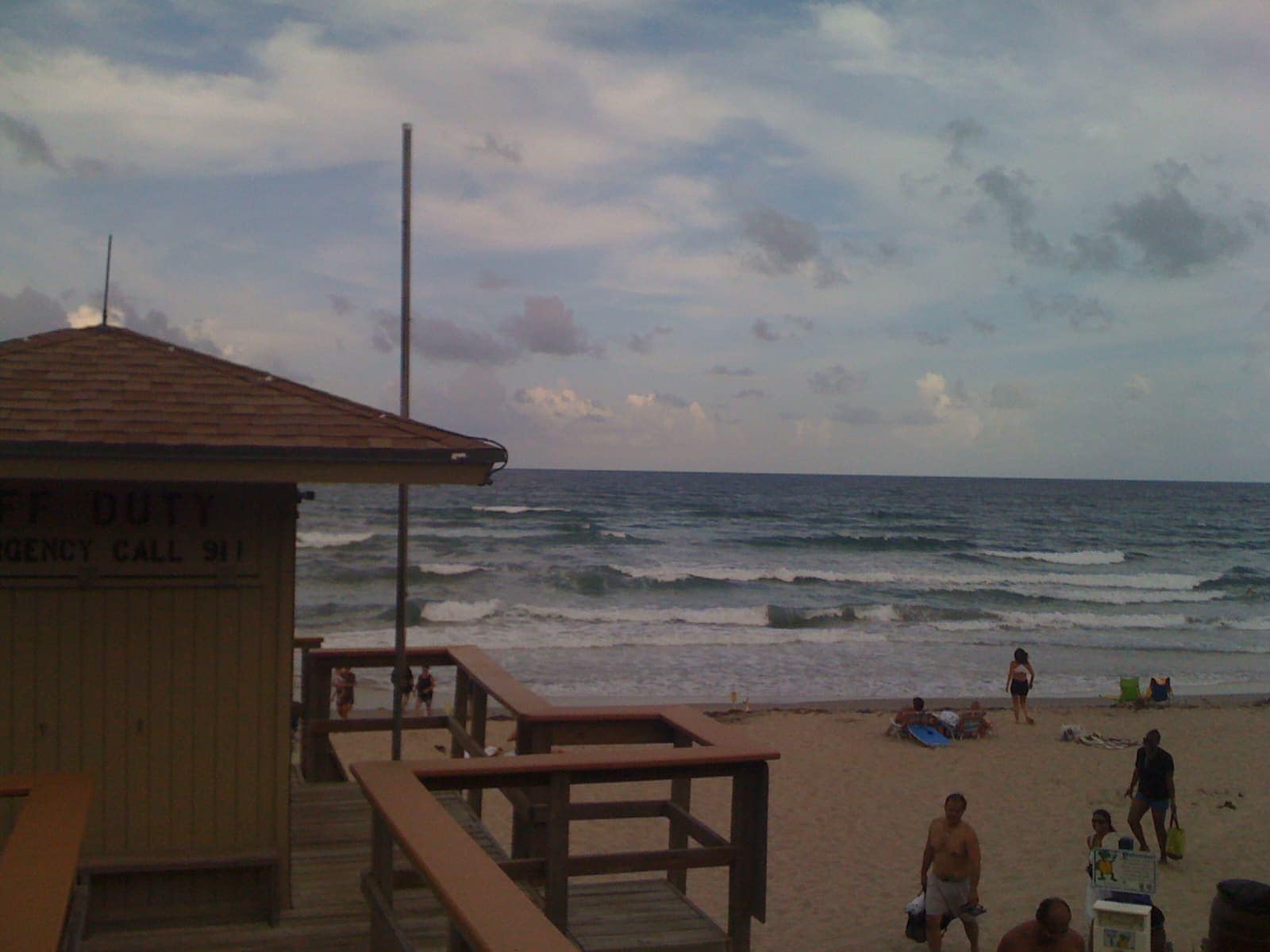 A sandy beach with a lifeguard stand, small distant people and rolling ocean waves under a cloudy sky.