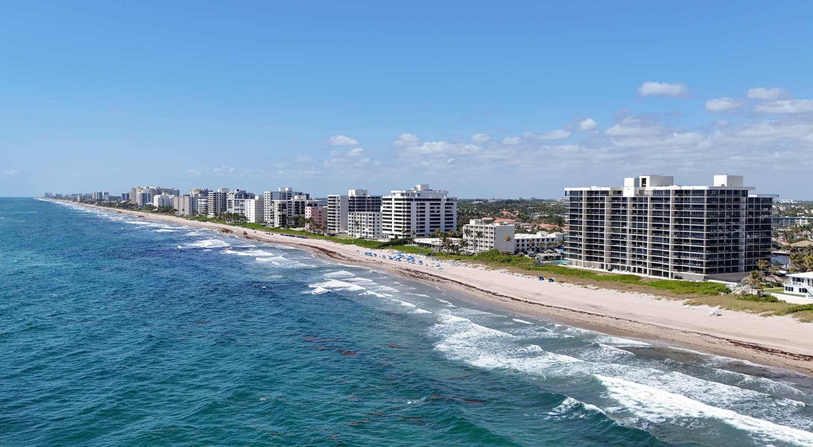Aerial view of sandy beach, turquoise surf and tall condominium towers under blue sky.