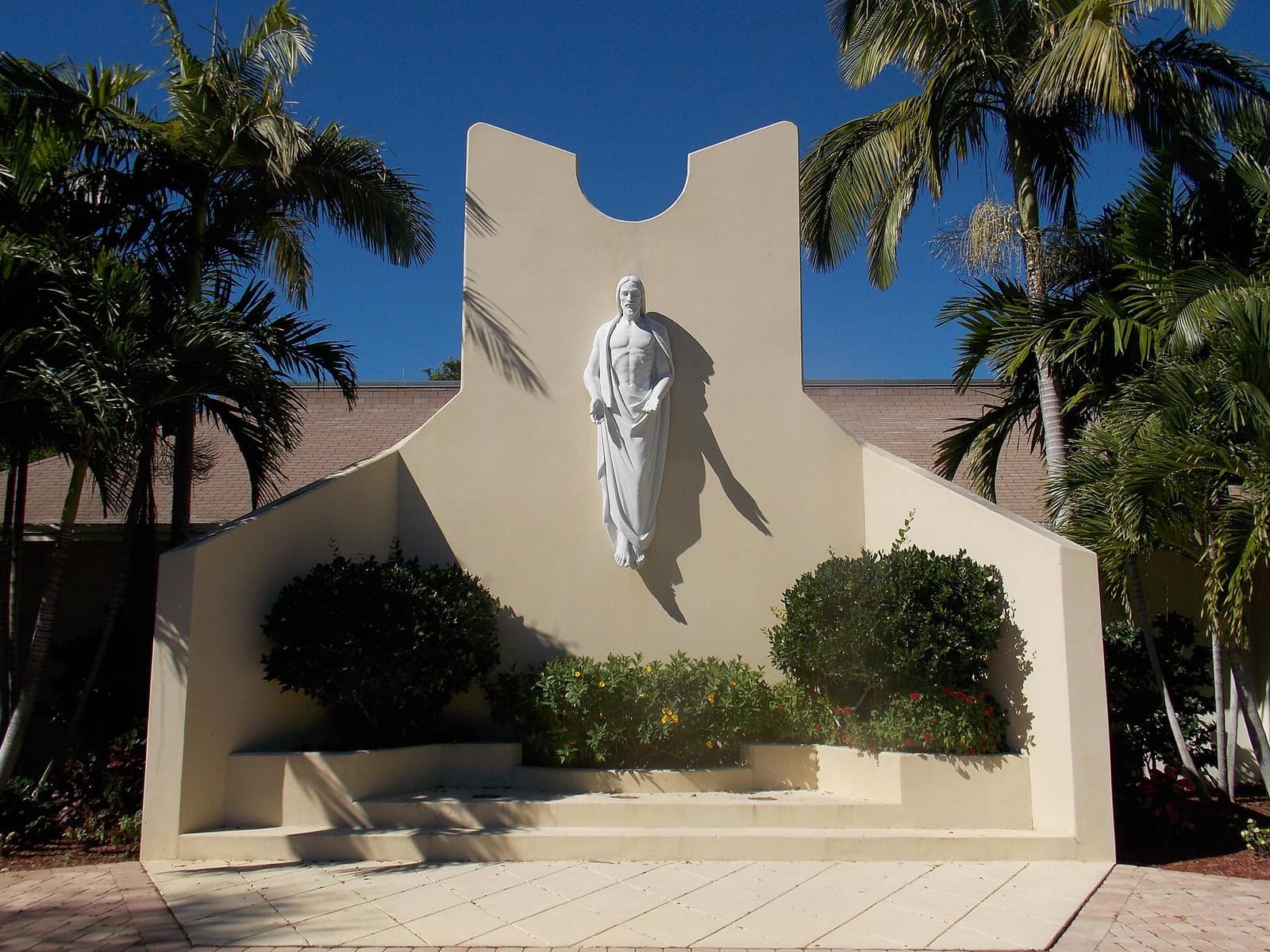 White statue of a robed figure on a beige wall framed by palm trees under a blue sky.
