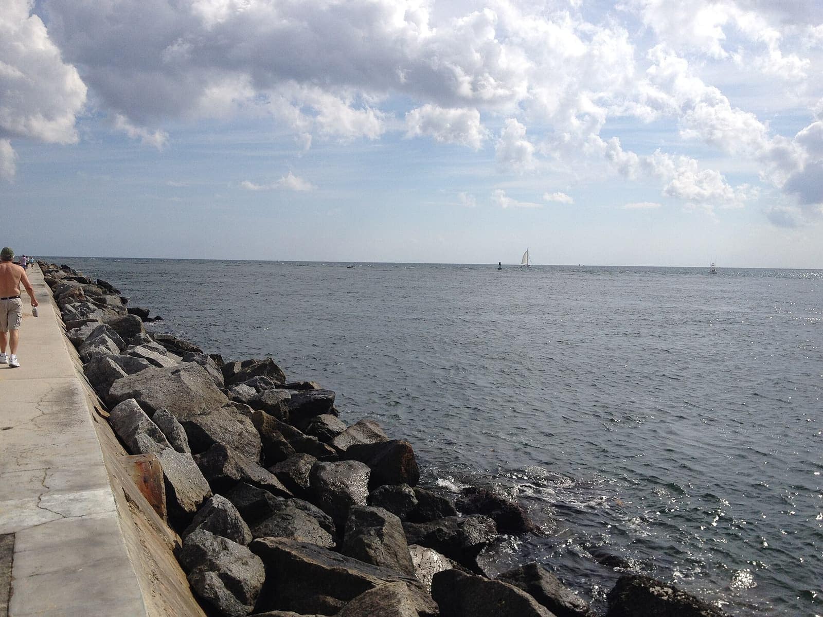 Rock jetty runs along a concrete walkway toward open ocean, with a sailboat and a distant walker.