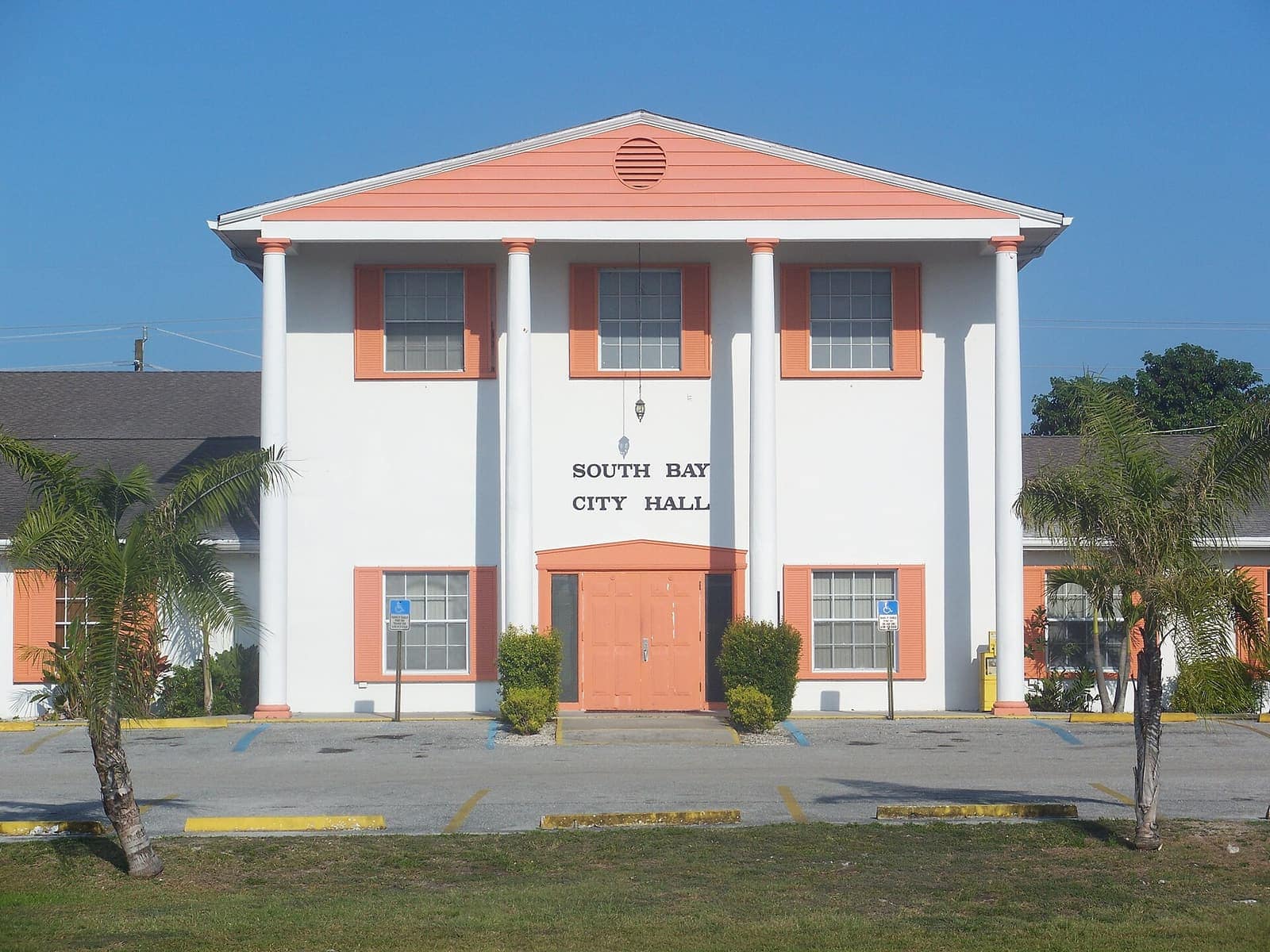 Two-story white city hall with peach trim and palm trees under a blue sky.