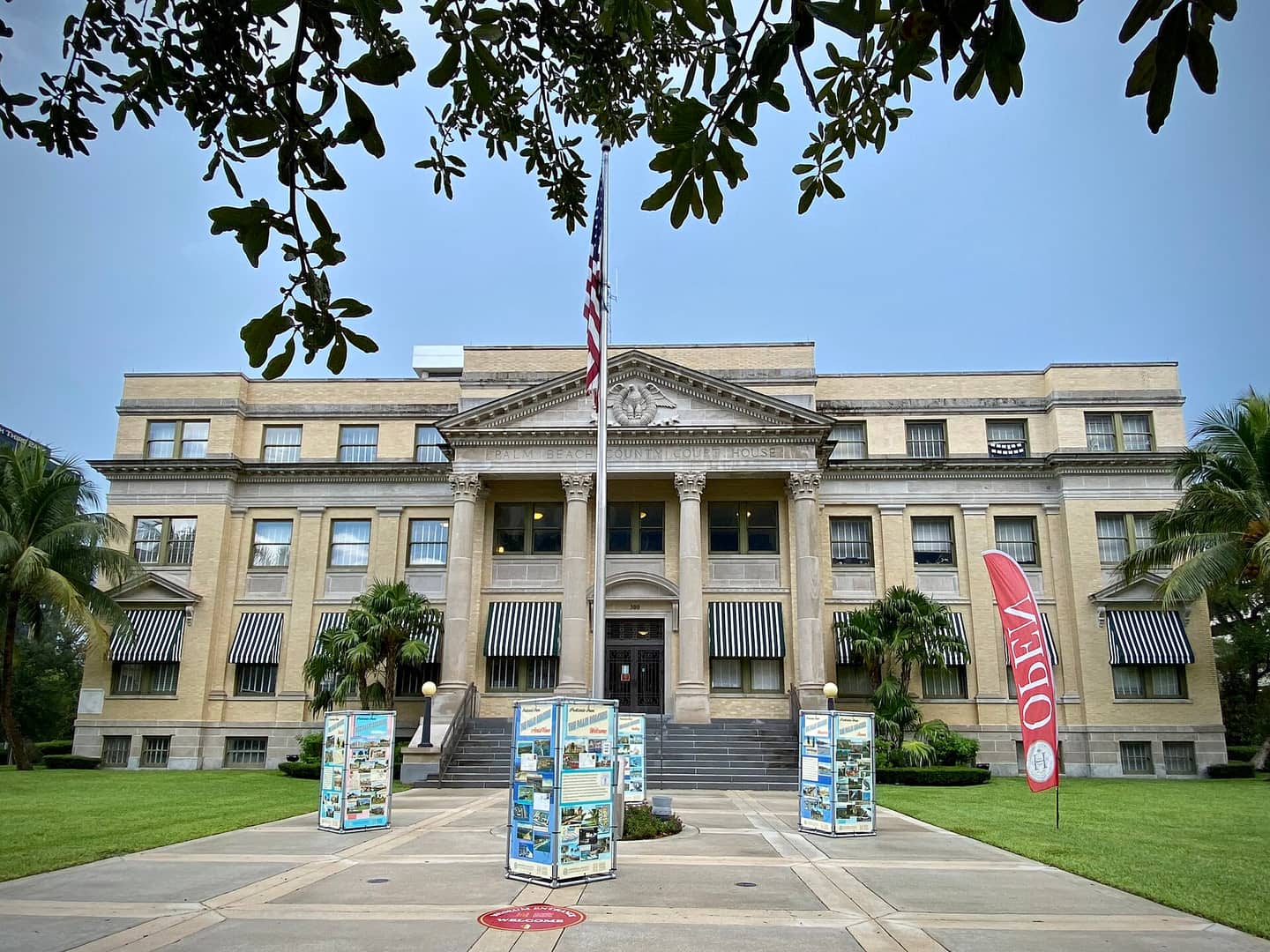 Tan stone courthouse with columns and a flagpole, framed by palm trees and tourist brochure stands.