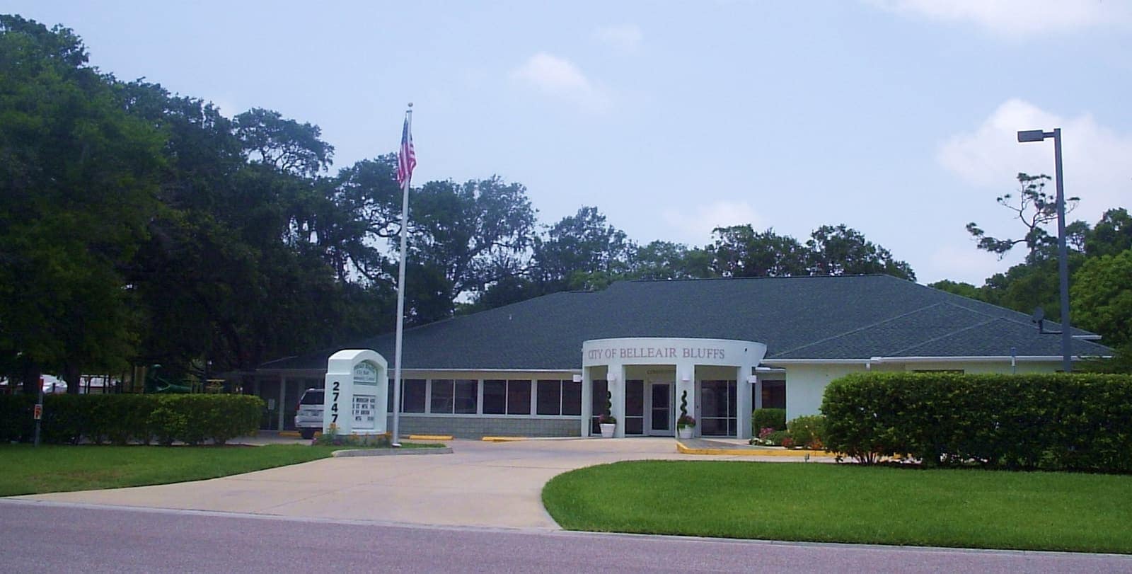 Single-storey white civic building with a flagpole and lawn beneath tall green trees on a sunny day.