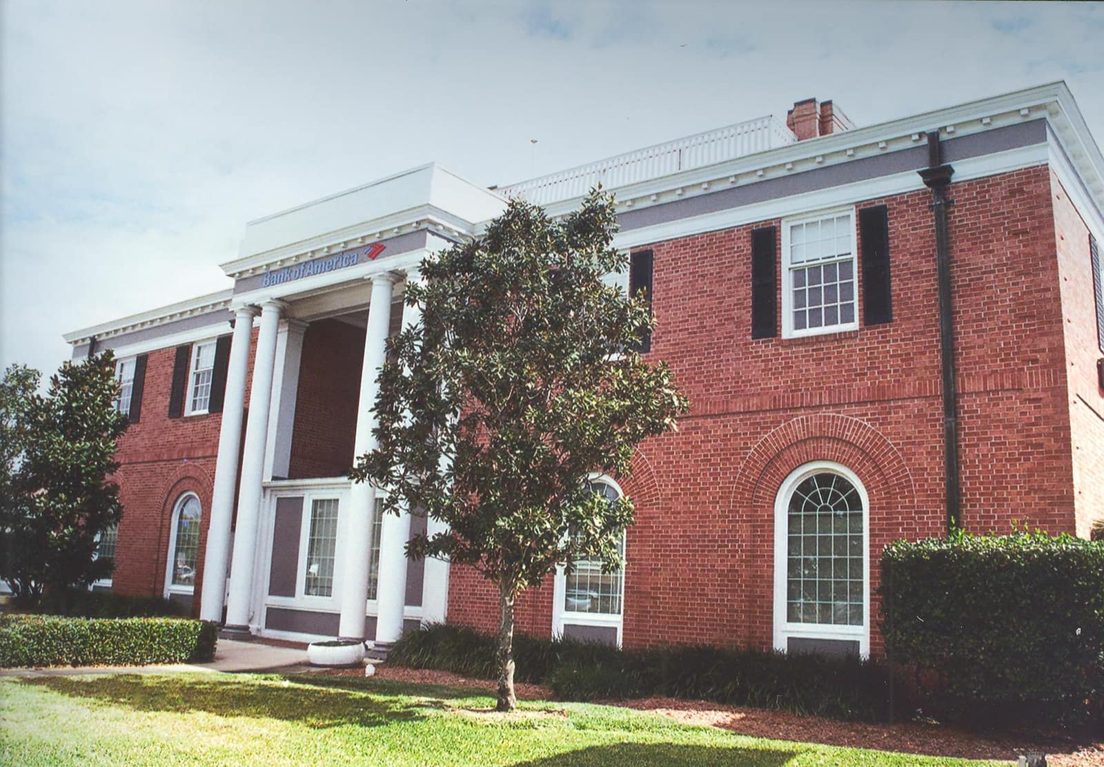 Two-storey red brick building with white columns, a young tree and green lawn under a pale sky.