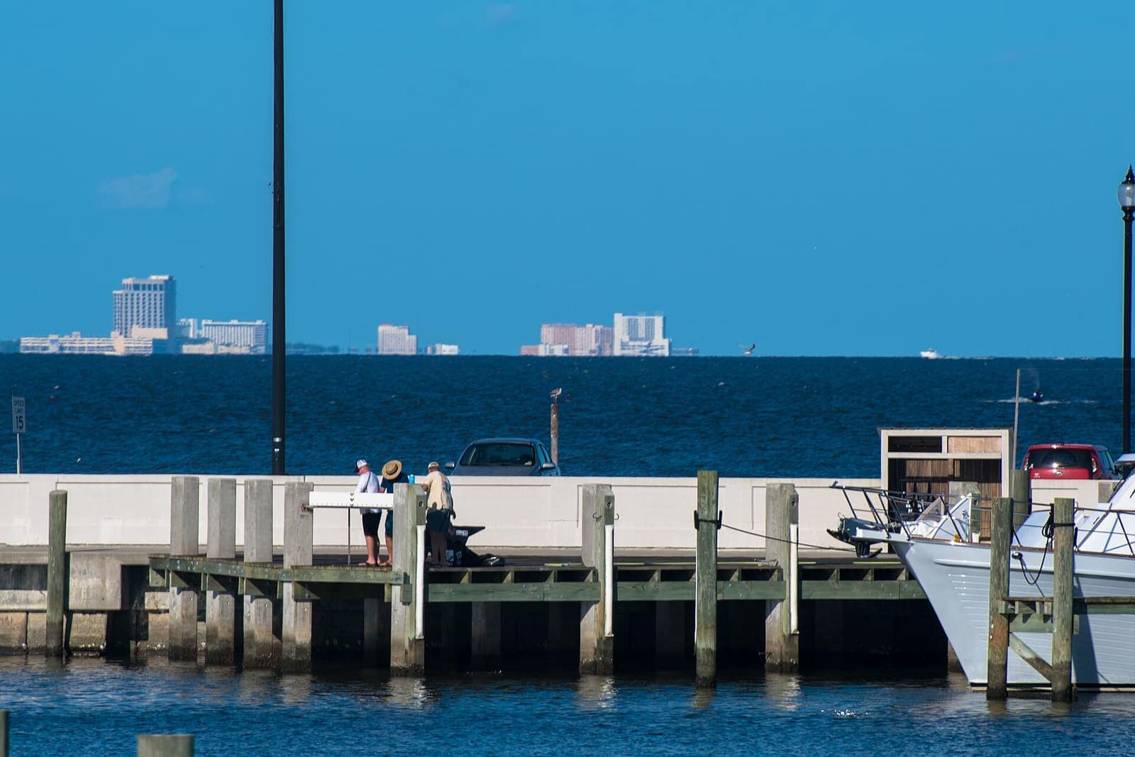 A concrete fishing pier over calm blue water with small distant people, moored boats and a hazy city skyline.