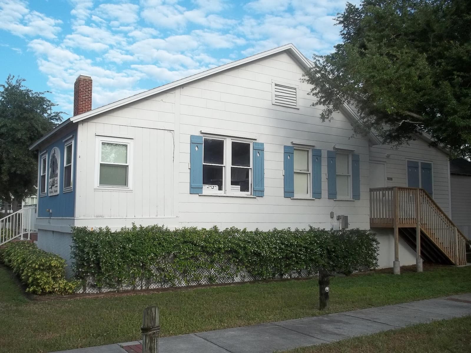 White wood-sided cottage with blue shutters, a brick chimney, and a green hedge along the lawn.