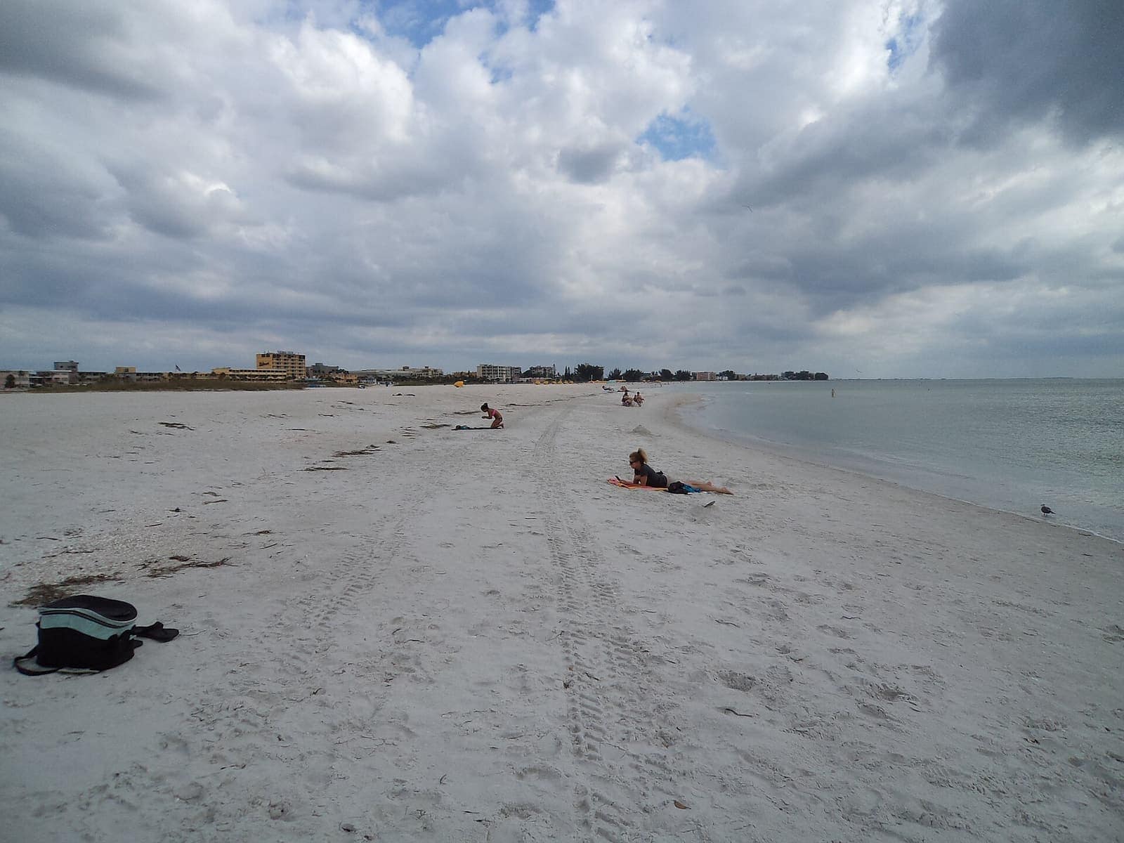 Wide white sand beach under cloudy sky, with small distant sunbathers and calm water on the right.