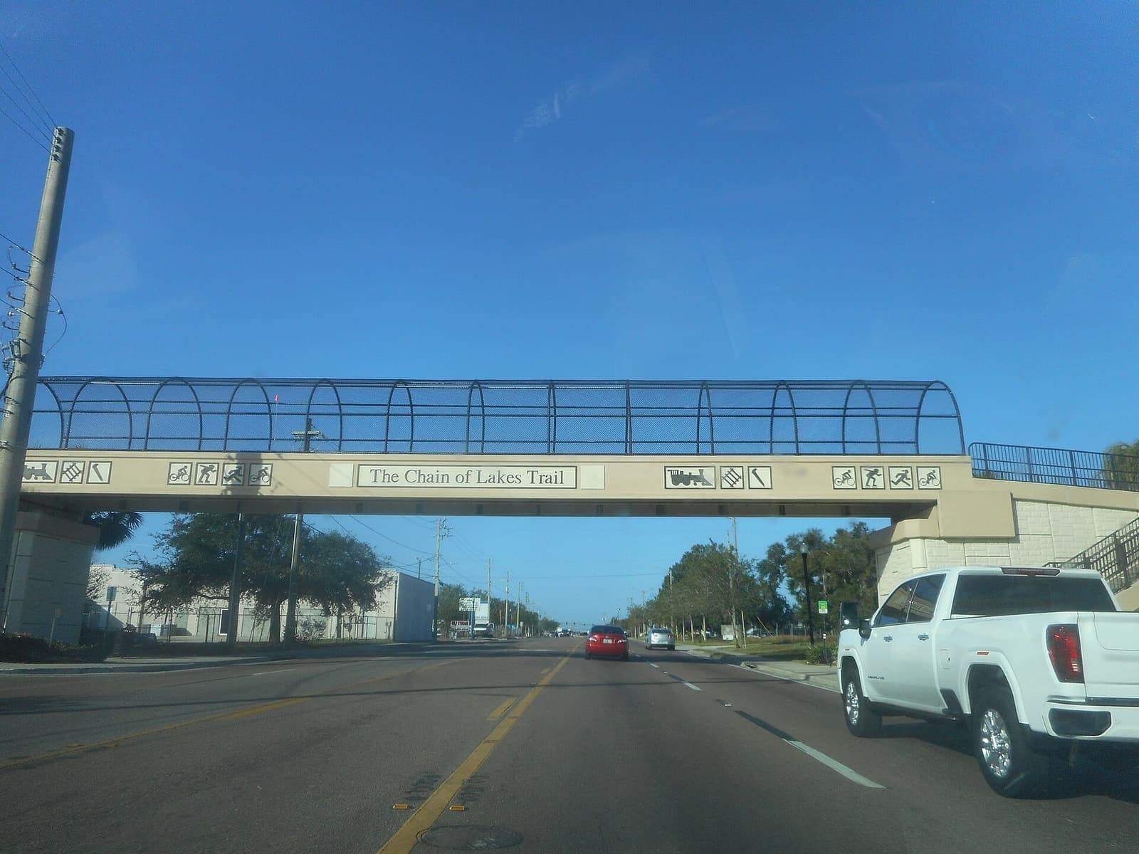 A pedestrian overpass labeled The Chain of Lakes Trail spans a wide road under a clear blue sky.