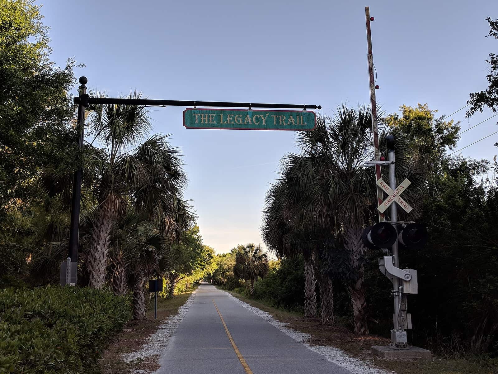 A paved trail runs beneath an overhead green sign reading The Legacy Trail, lined with palms and trees.