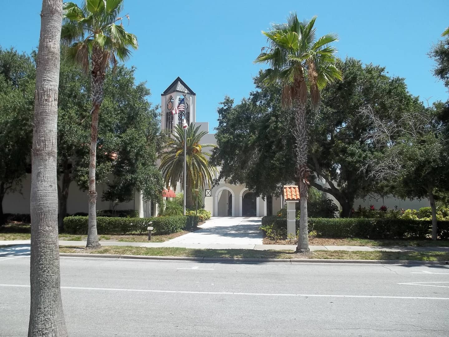 White Mediterranean-style civic building with a bell tower behind tall palm trees beside a wide street.