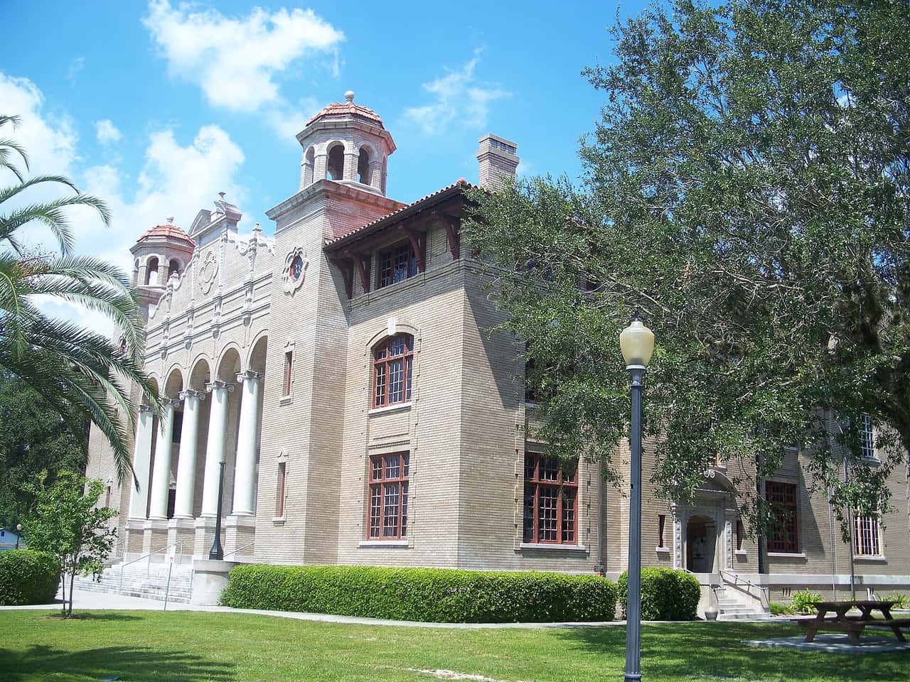 Historic brick courthouse with arched colonnade and towers, framed by a large oak tree and palm.