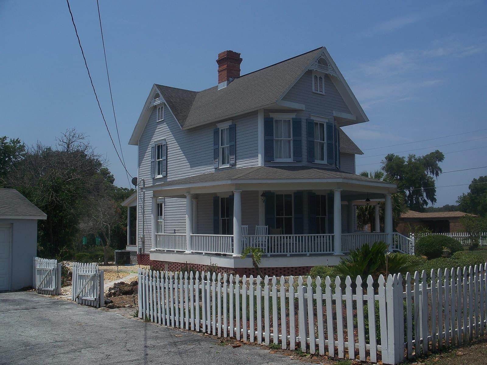 Two-story white house with blue shutters, wraparound porch and white picket fence under blue sky.