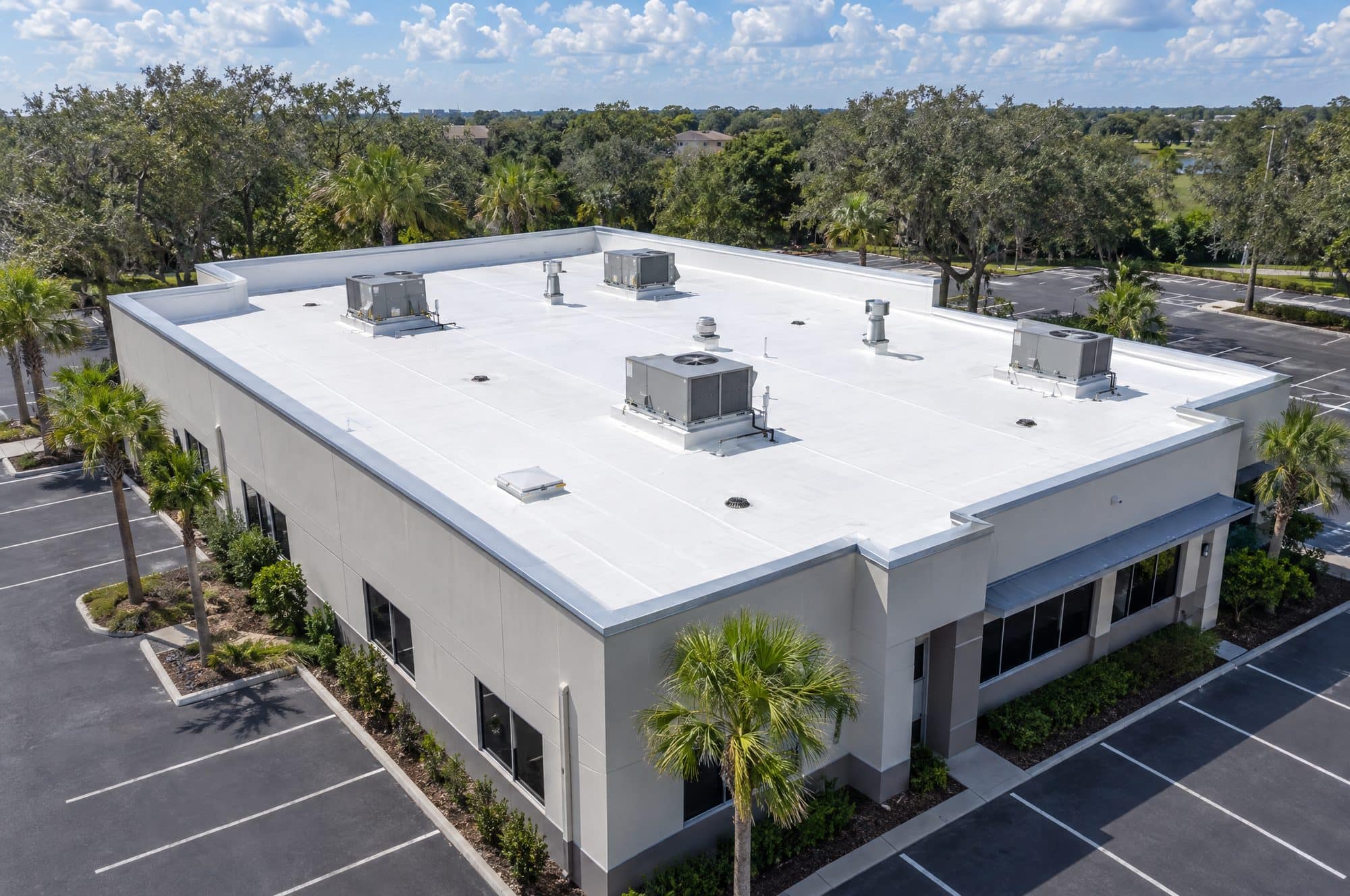 Drone view of a single-story commercial building with a white TPO flat roof, parapet with metal coping and rooftop HVAC units, empty parking lot and live oaks (illustration)