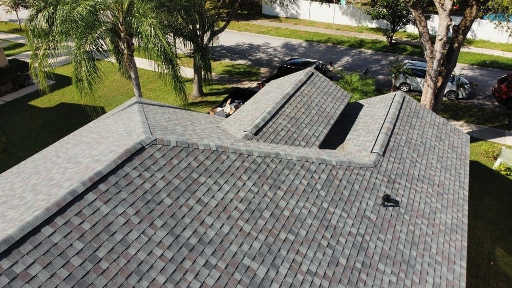 Low aerial view over a gray-blend shingle roof with gables and valleys, palm trees, a street and parked cars behind