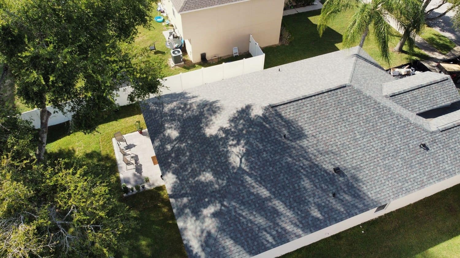 Drone view looking down on a single-story home with a gray shingle roof, a backyard patio with chairs, a white vinyl fence and palm trees.