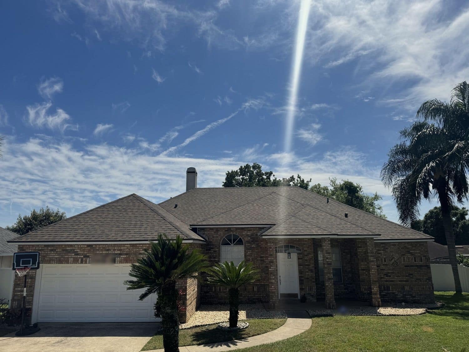 Front of a single-story brick home with a dark shingle hip roof, a white garage door and two small palms in the front bed