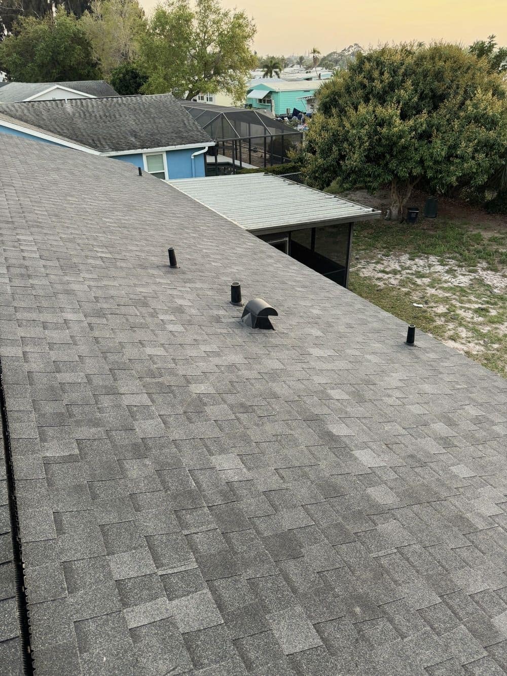 View down a gray shingle roof slope with pipe boots and a roof vent, neighboring homes and a screened pool enclosure below.