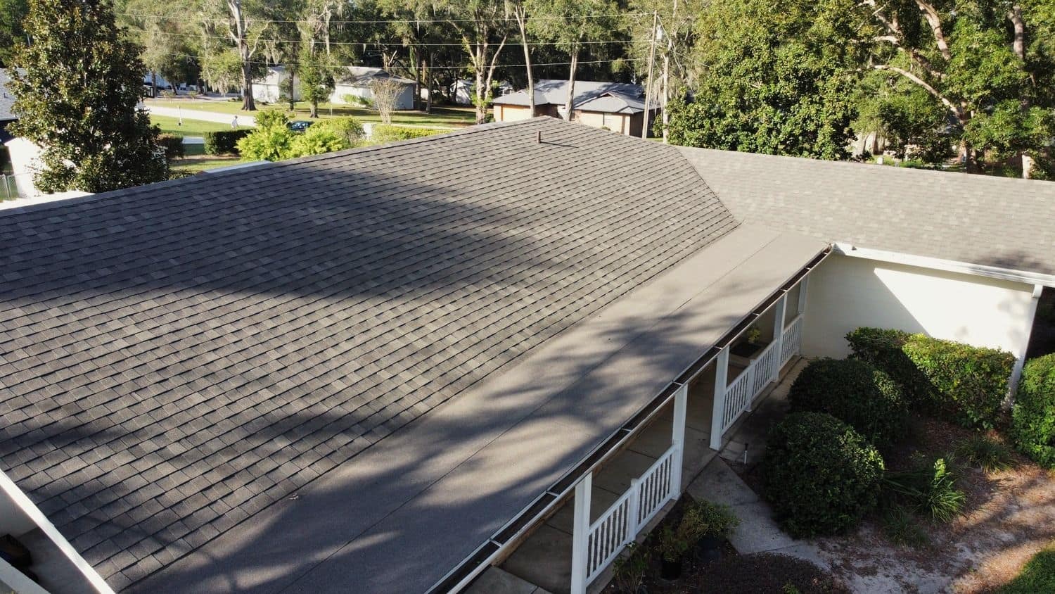 Elevated view across a gray shingle roof above a covered porch with white railings, shrubs along the walk and tall trees behind.